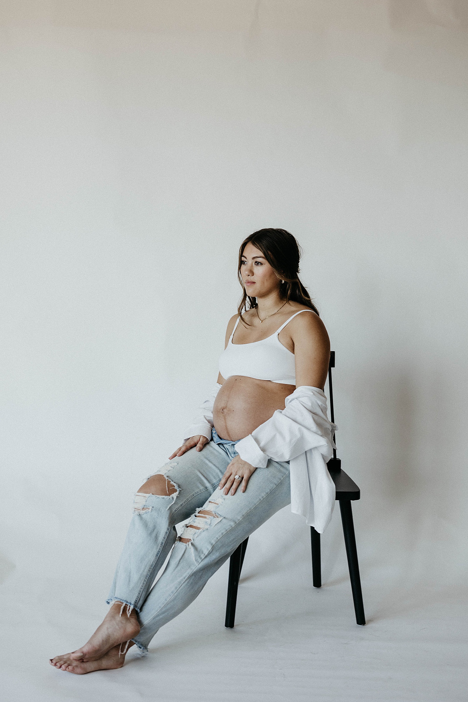 A pregnant woman sits on a stool against a white backdrop in a studio maternity photoshoot.