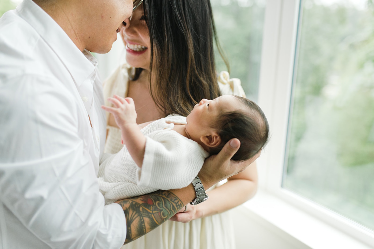 A mother and father smile and hold their newborn beside a window.