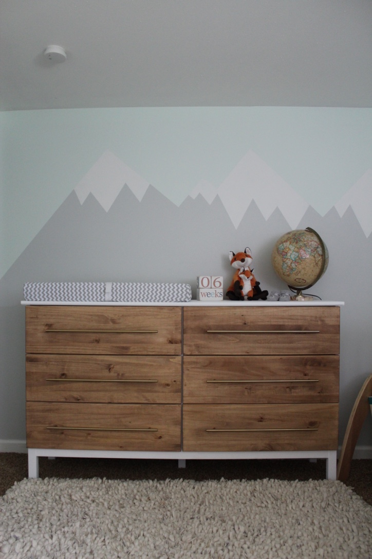 An Ikea dresser sits in front of a mountain mural and forest themed nursery decor.