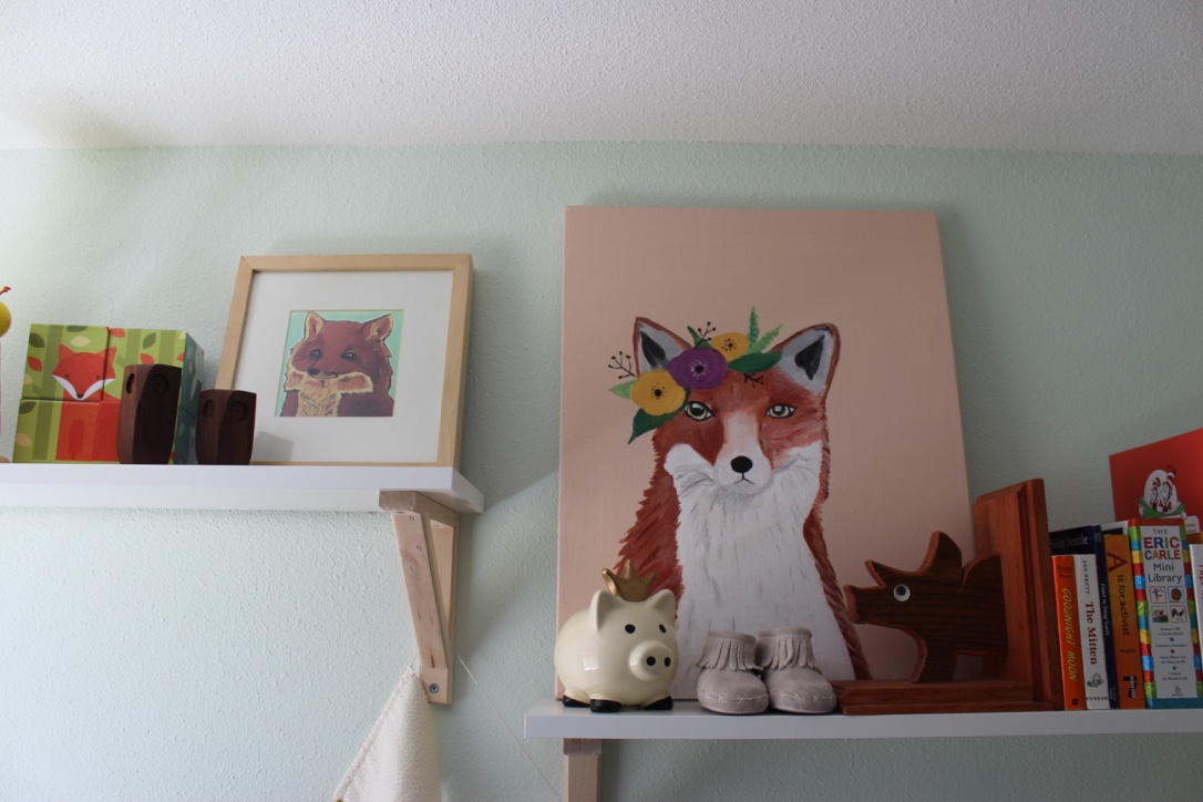 Paintings of foxes sit beside books on white shelves in a baby's nursery.