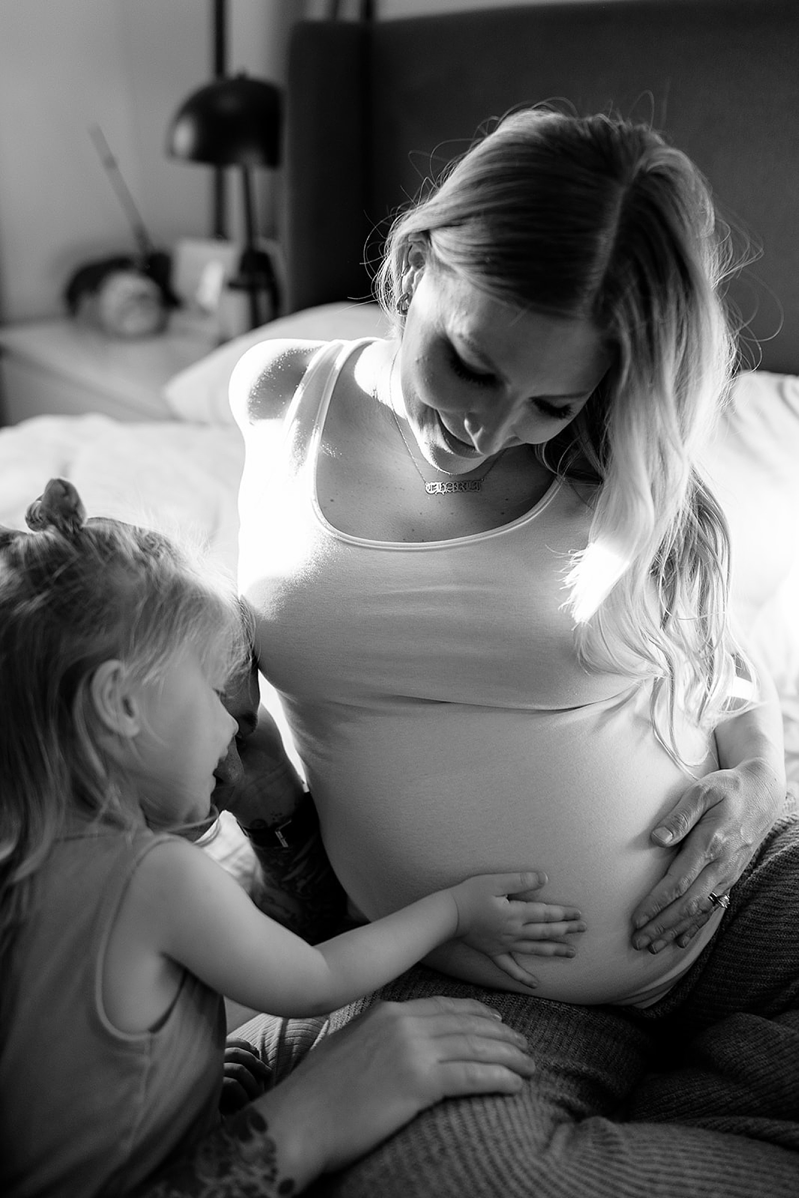 A toddler places her hand on her mother's baby bump while they sit on a bed.